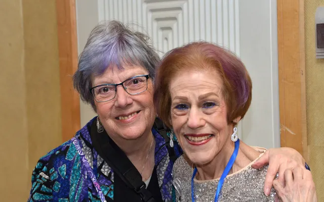 Two elderly women smiling, embracing at an event.