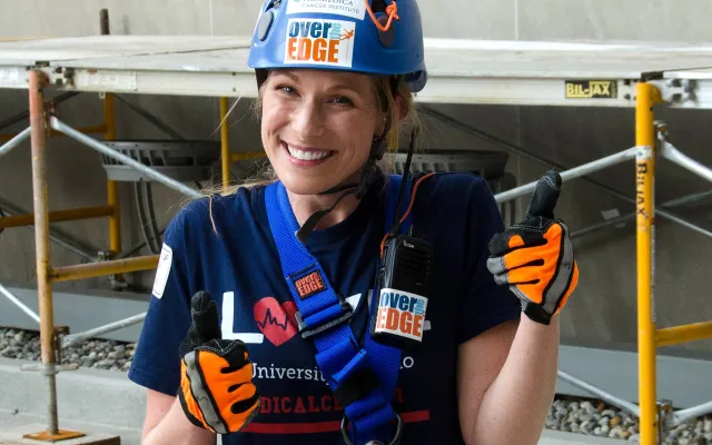 A person in climbing gear smiling and giving thumbs up indoors.