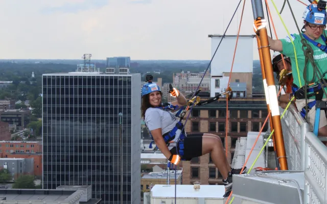 Person rappelling down a tall building in an urban setting with safety gear.