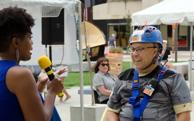Woman with microphone interviews man in climbing gear.