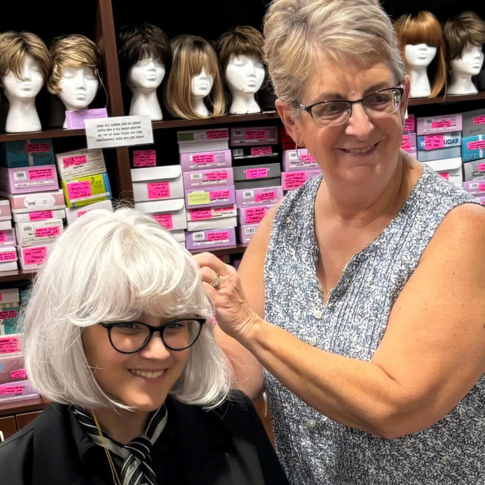 Two women smiling in a wig store with numerous wigs on shelves.