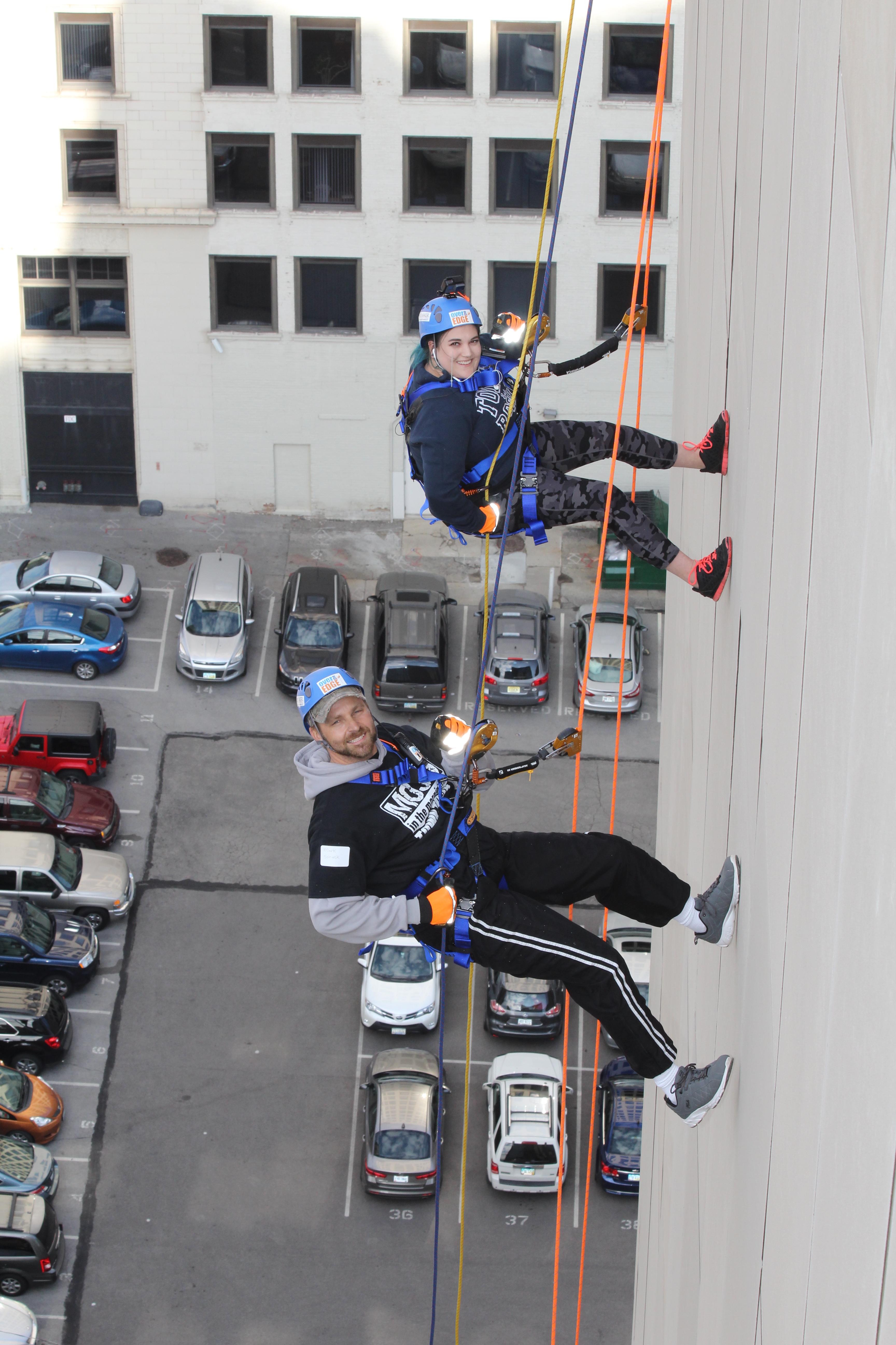 Two people rappelling down a tall building, smiling, parking lot below.