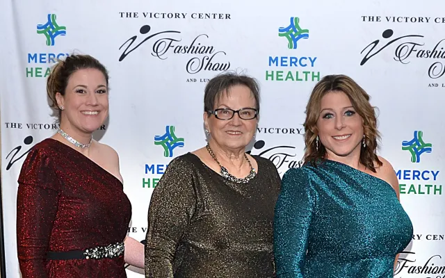 Three women in evening gowns stand smiling in front of a Mercy Health backdrop.