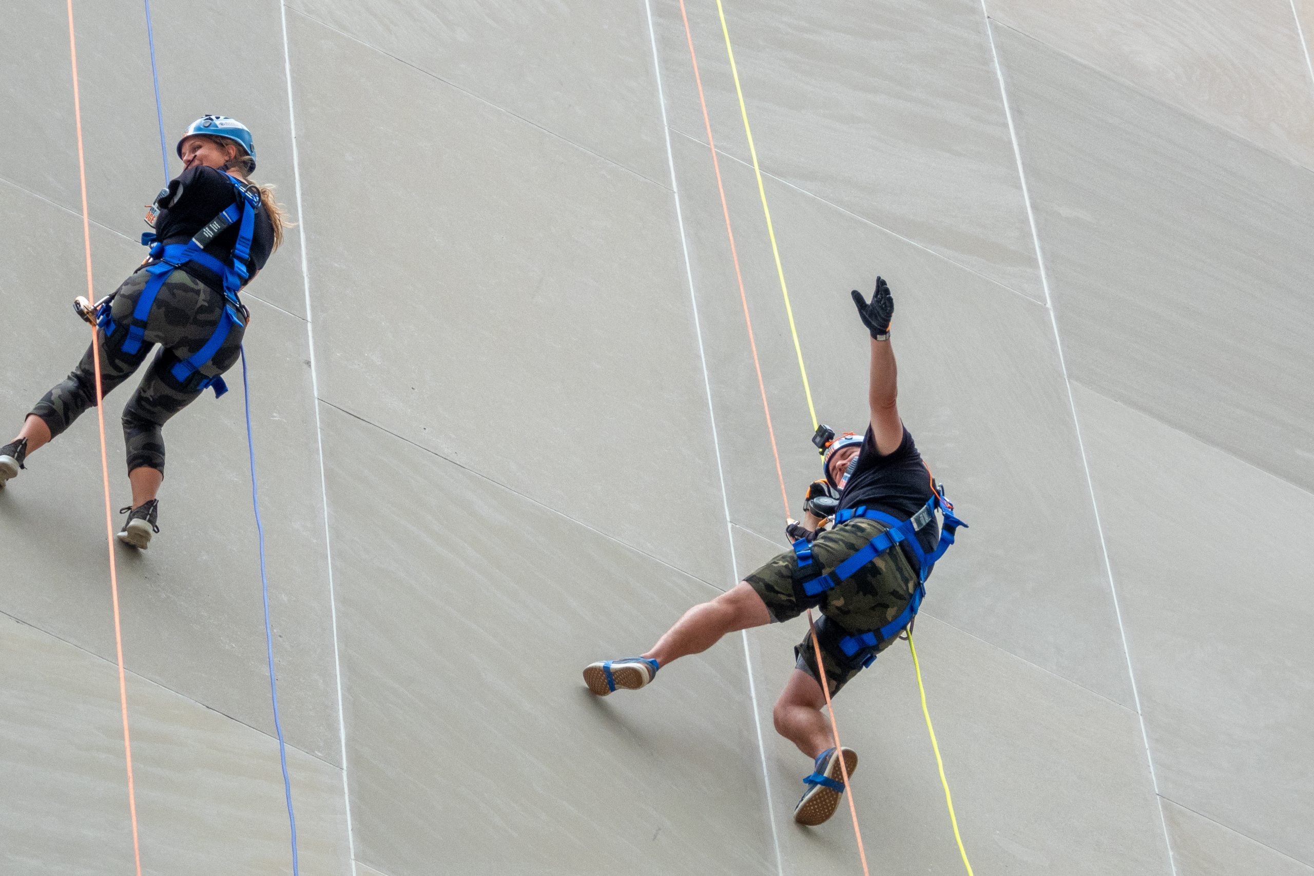 Two people rappel down a gray towering wall with colorful ropes.