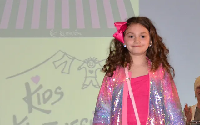 Young girl in pink and sparkly outfit walks on runway, smiling.