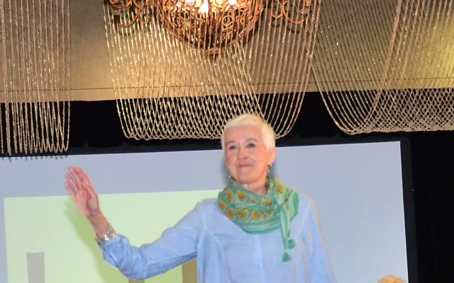 Senior woman models on a runway, wearing light blue and white clothes with a colorful scarf.