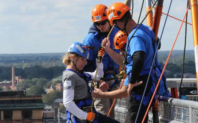 Three people in helmets and harnesses prepare to rappel down a building.