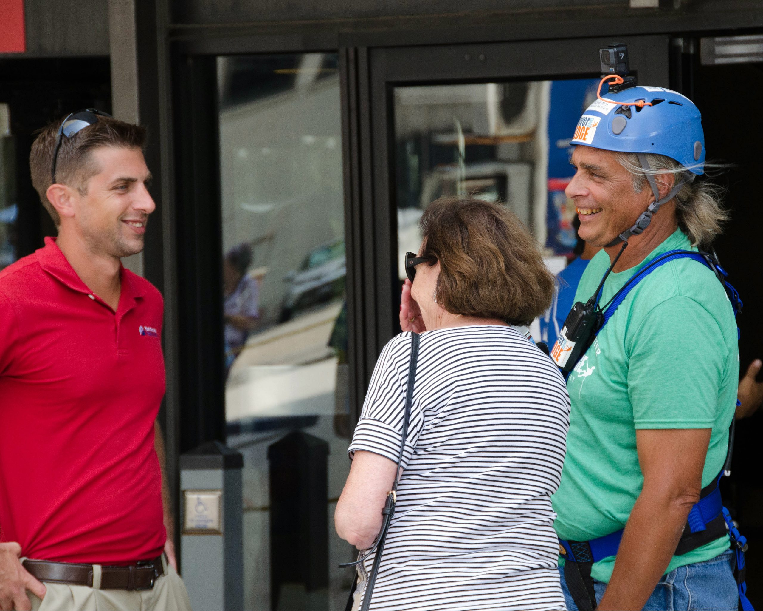 Three people chatting outdoors; one man wears a helmet and harness.
