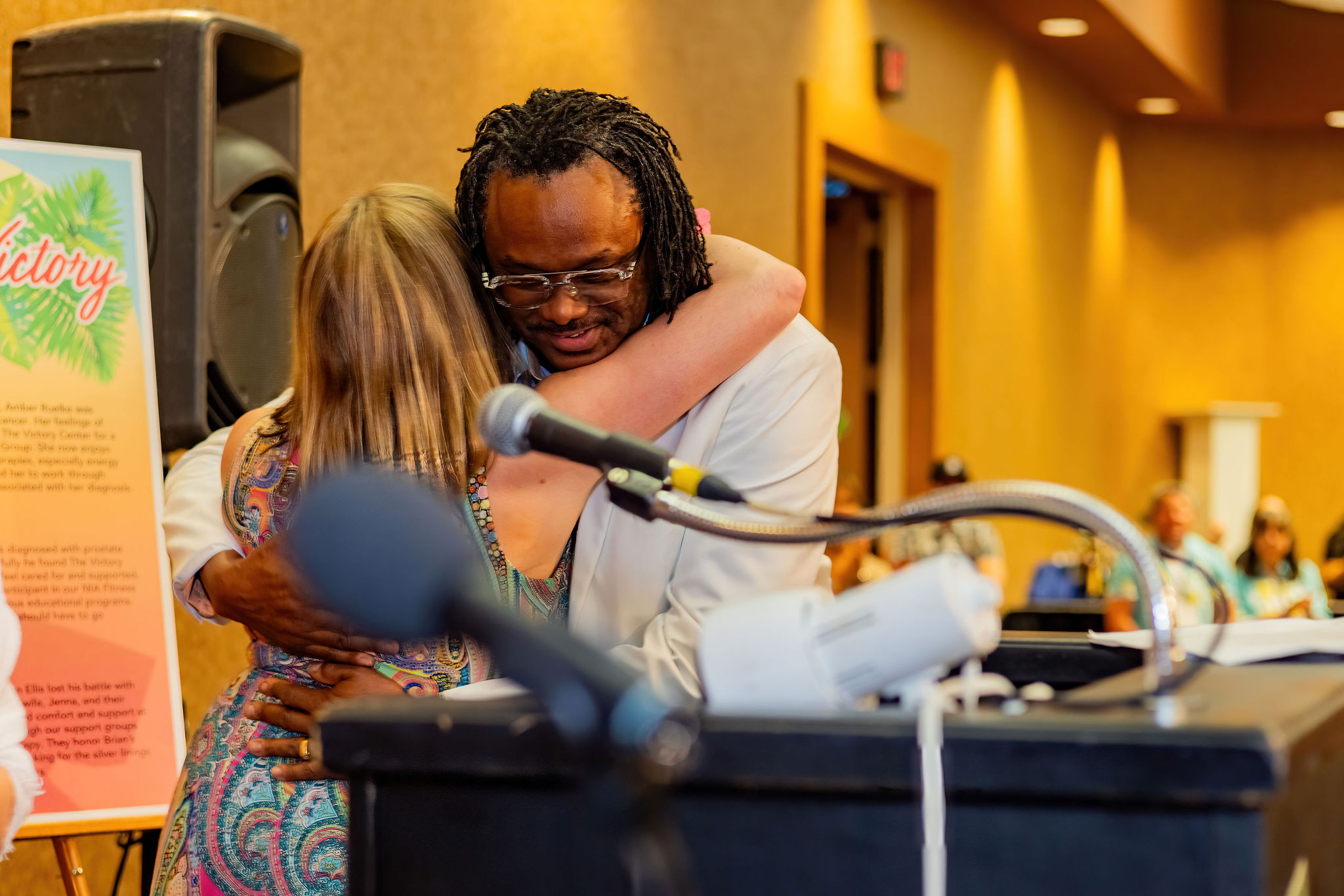 A man and woman hug warmly behind a podium in a conference room.