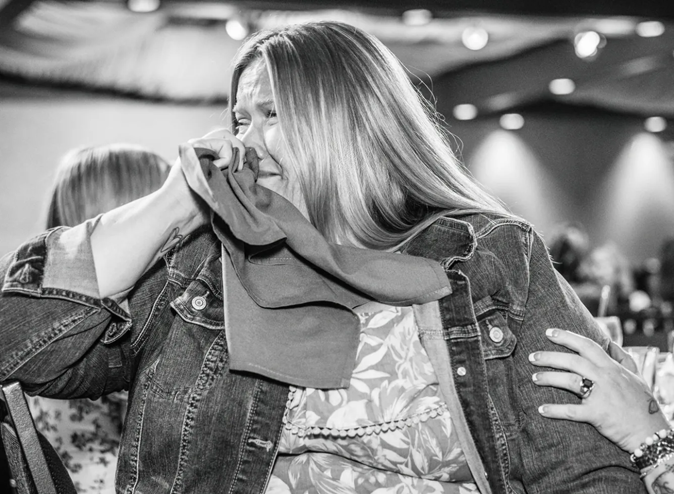 Woman in a denim jacket crying with a tissue, hand on her shoulder for comfort.