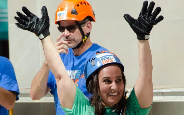 Woman in climbing gear excitedly raising arms, man in background wearing helmet.