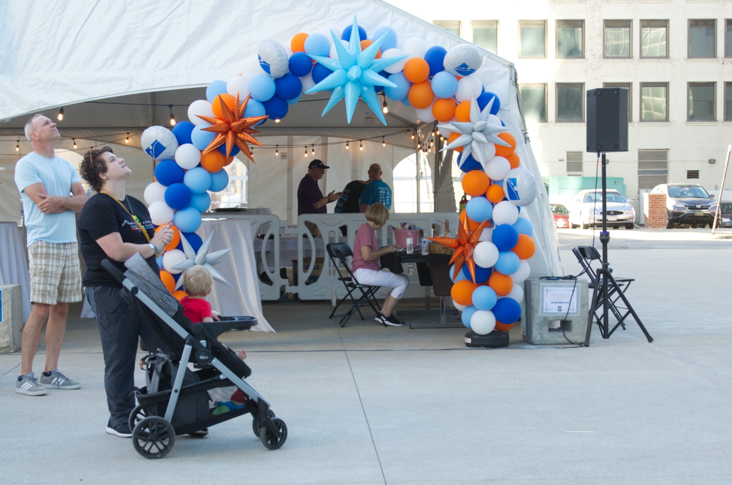 White tent entrance with colorful balloon arch; people standing and sitting outside.