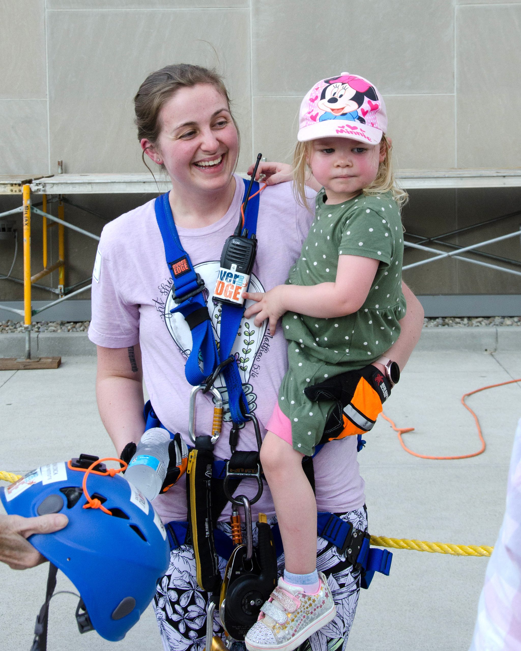 Woman in climbing gear holds a child wearing a Minnie Mouse hat, both smiling outdoors.