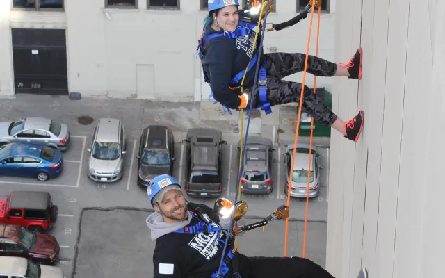 Two people rappelling down a tall building, smiling, parking lot below.