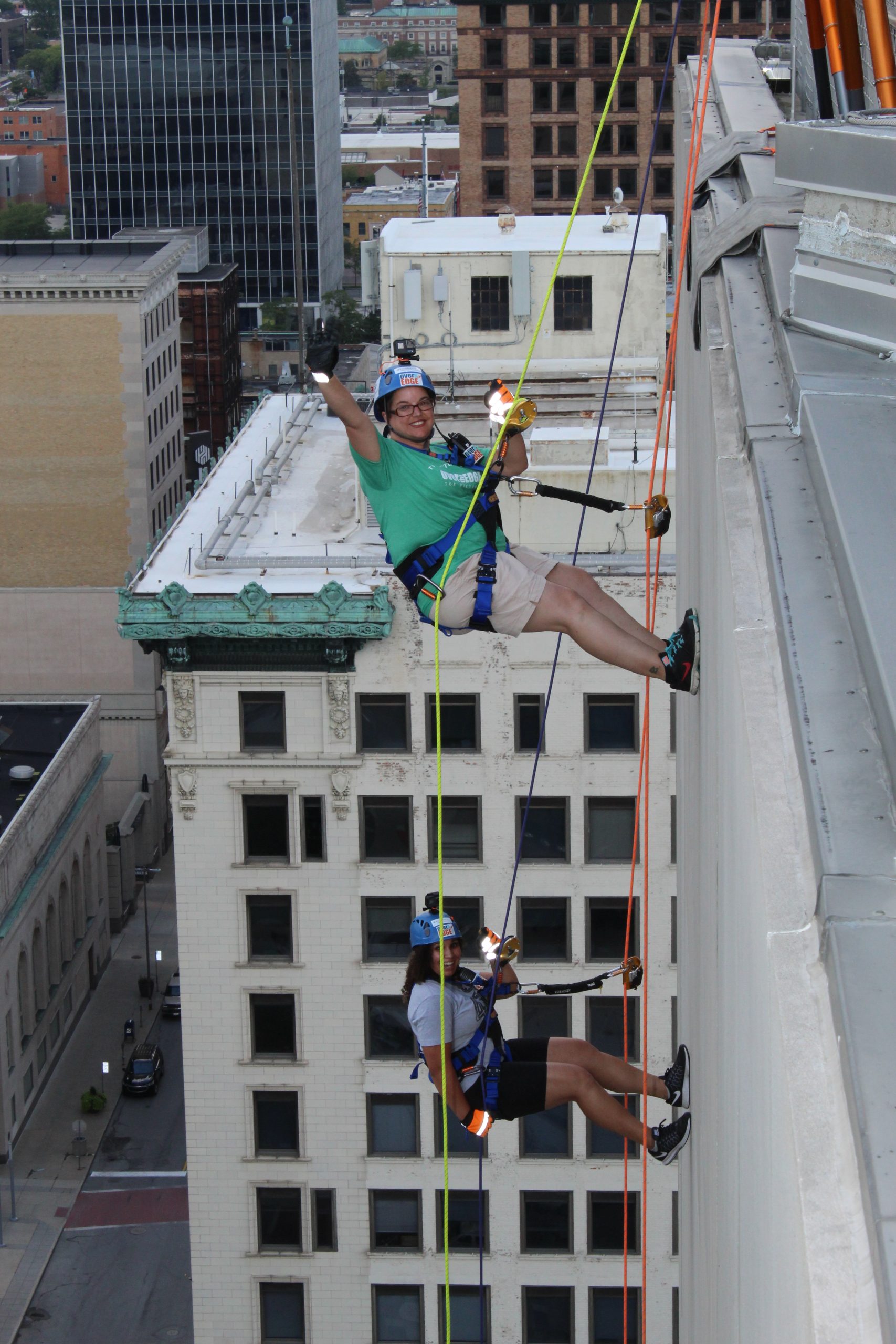 Two people rappelling down a tall city building, wearing safety gear.