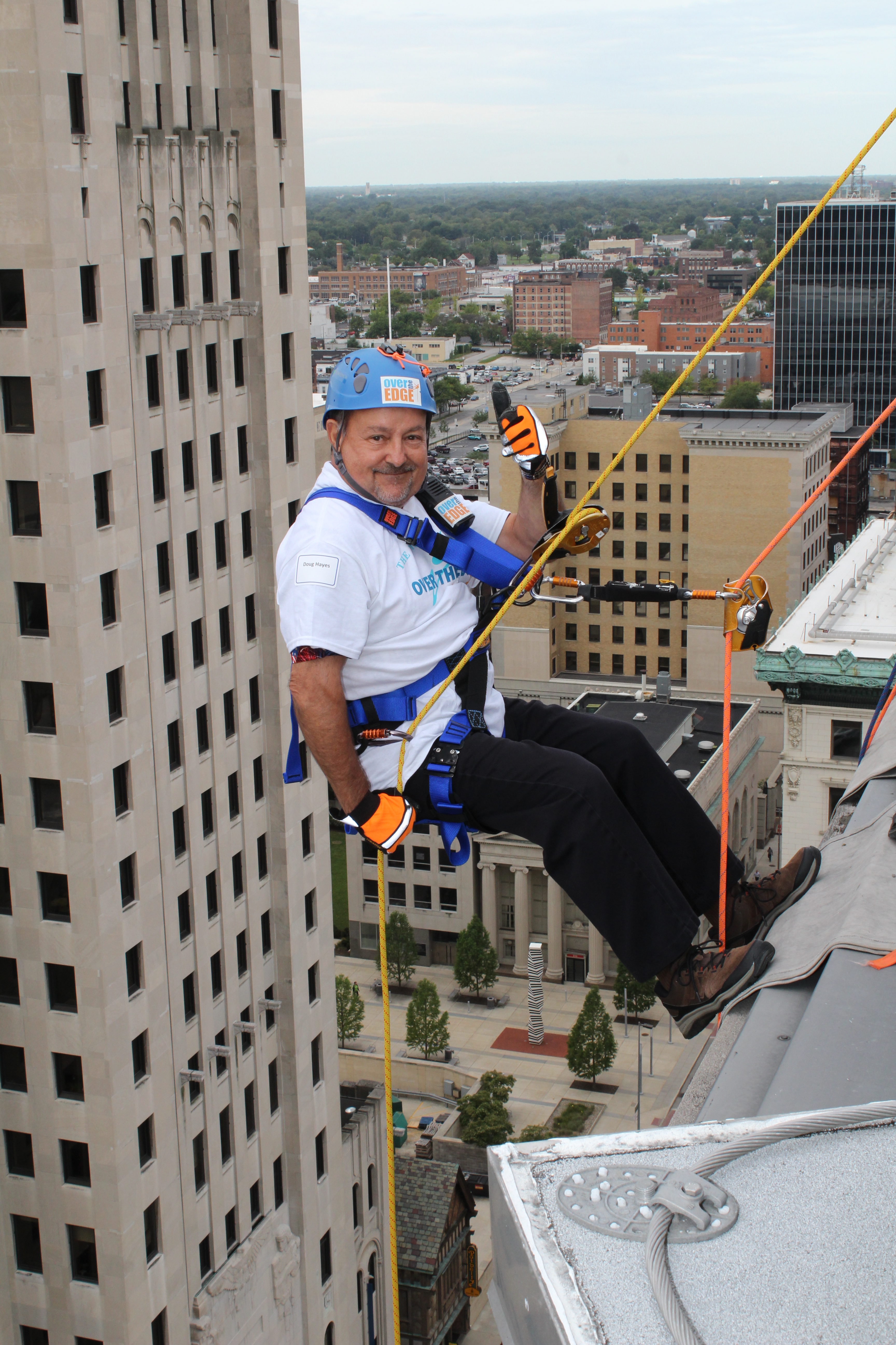 Person in safety gear rappels down a tall building, cityscape in the background.
