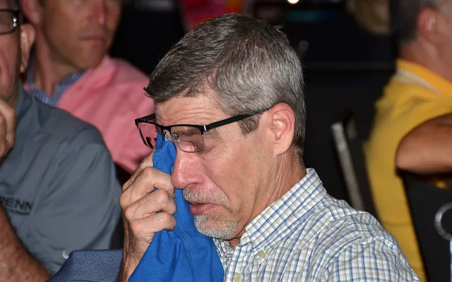 Man wearing glasses dabs eye with a blue cloth, surrounded by people.