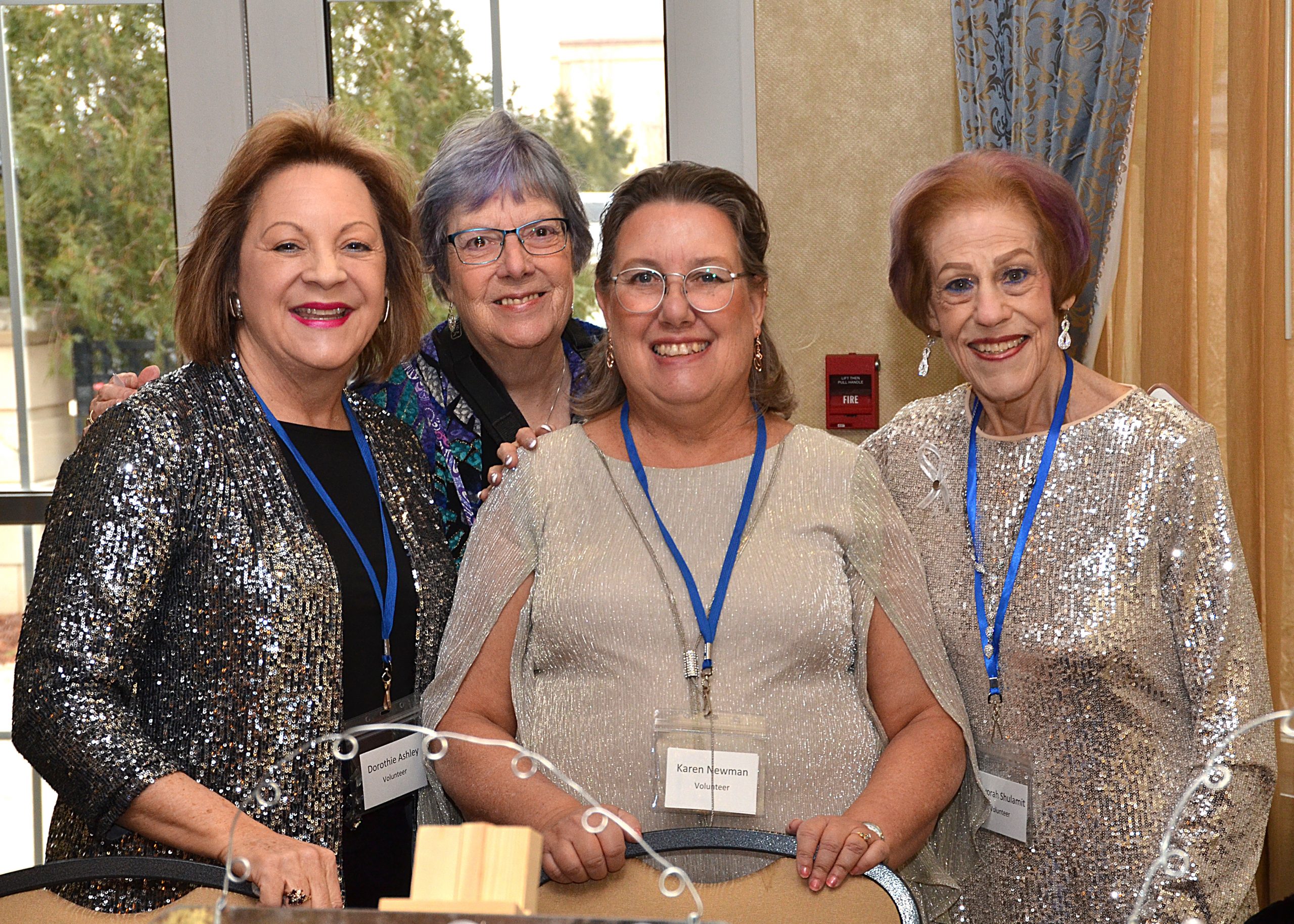 Four women smiling at an indoor event, wearing name tags and sparkly outfits.