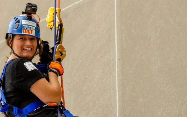 Person rappelling down a wall, wearing a helmet and harness, smiling.