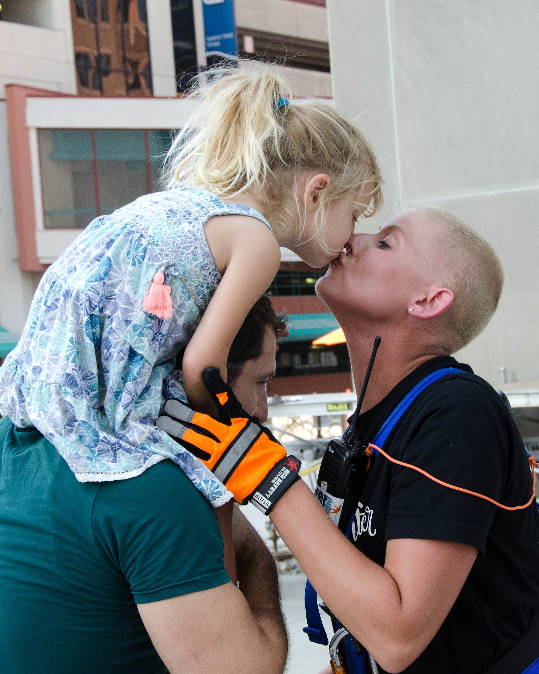 Child kissing adult, held up by another person, in a city setting.