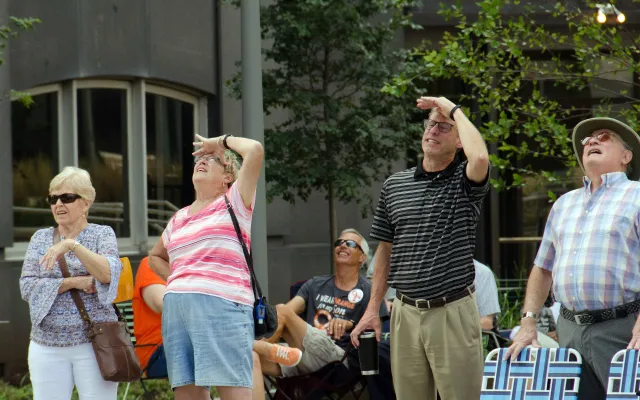 Four people looking up, shielding eyes from sunlight, with trees and buildings in the background.