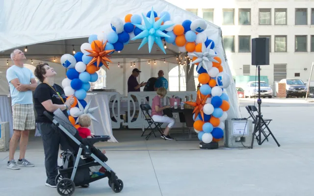 White tent entrance with colorful balloon arch; people standing and sitting outside.