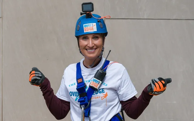 Person in climbing gear and helmet smiling at camera, urban setting.