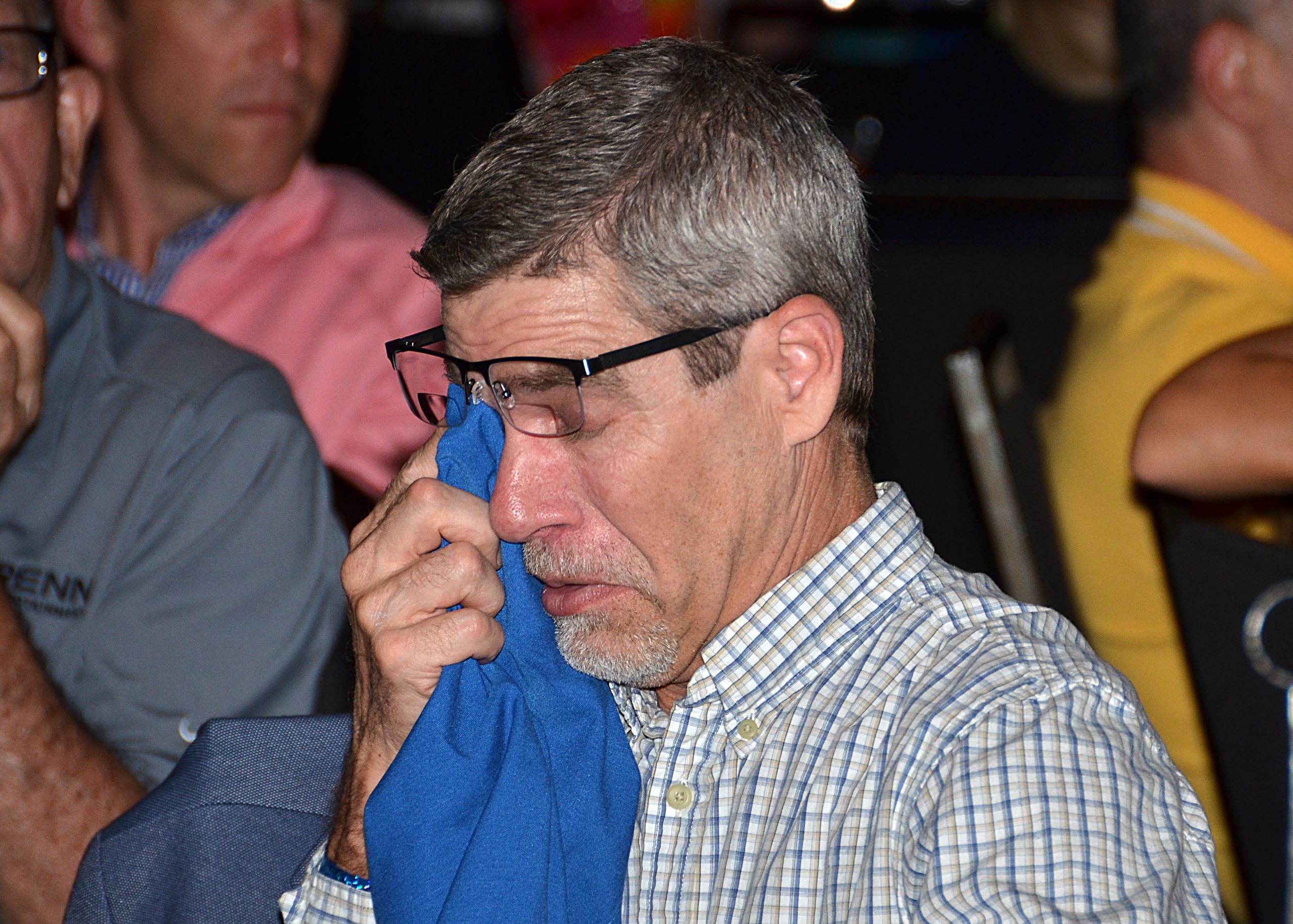 Man wearing glasses dabs eye with a blue cloth, surrounded by people.