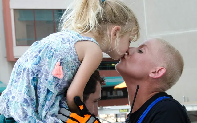 Child kissing adult, held up by another person, in a city setting.