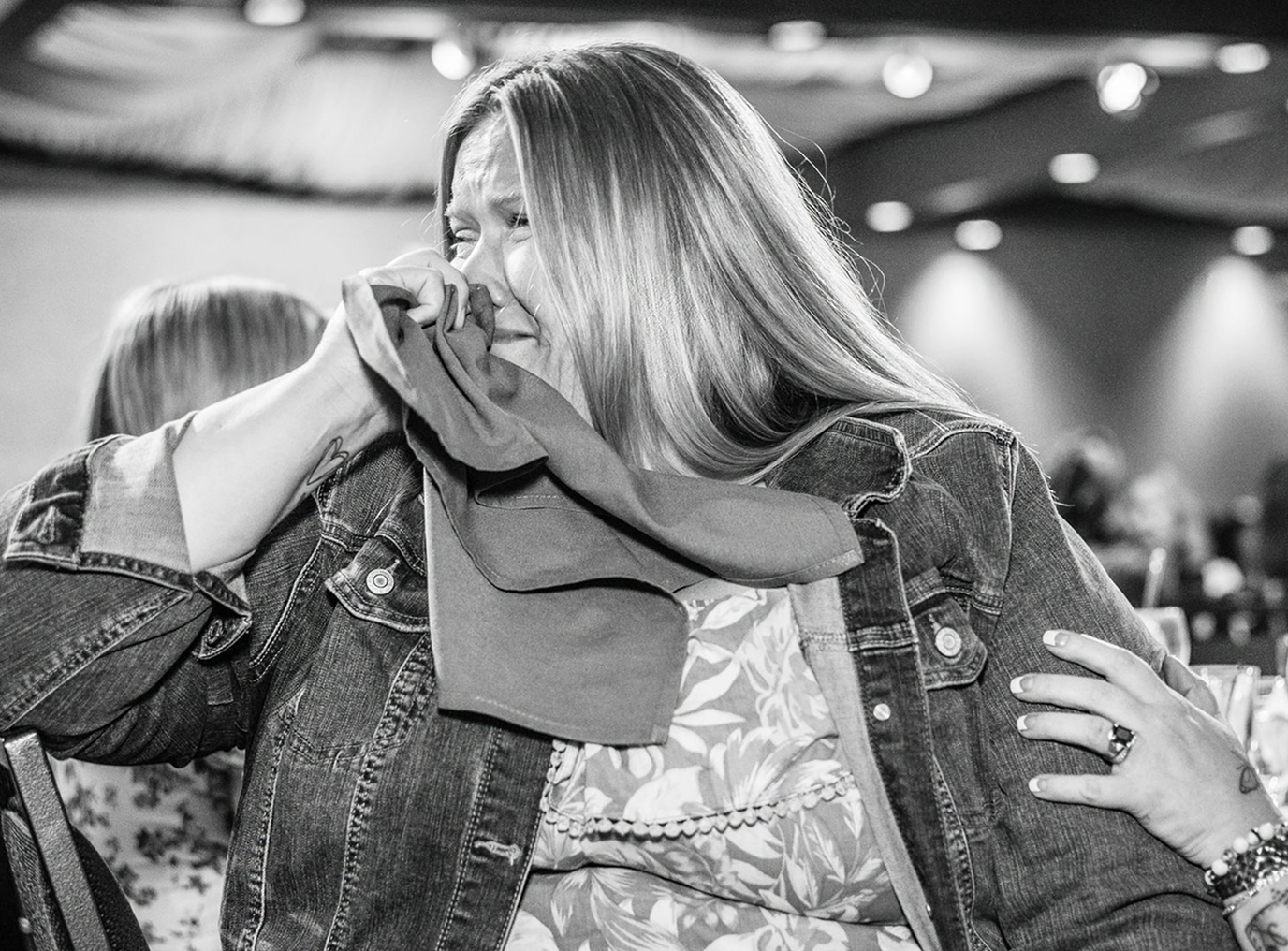 Woman in a denim jacket crying with a tissue, hand on her shoulder for comfort.