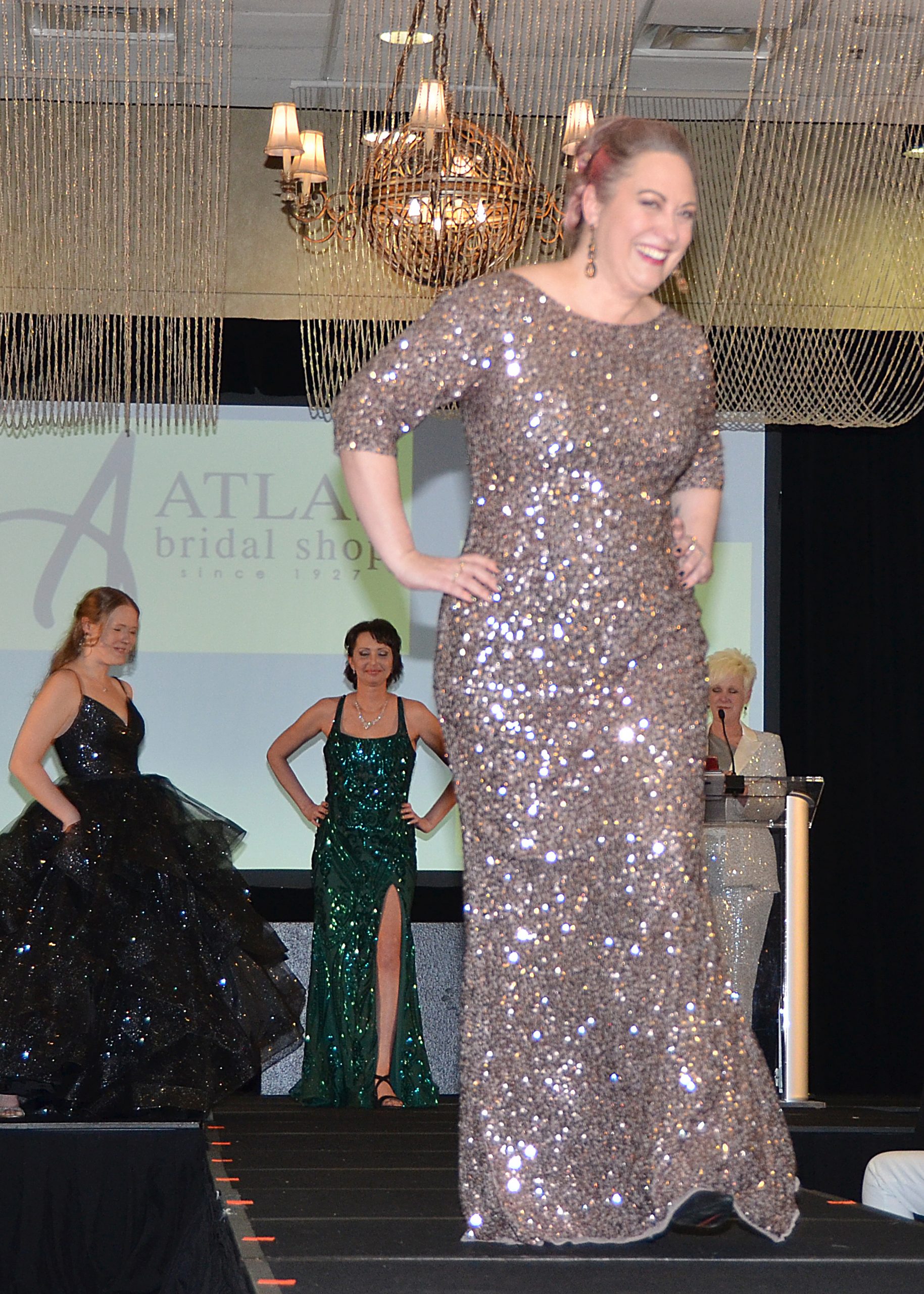 Model in a sparkling gown walks runway, others watch; chandelier overhead.