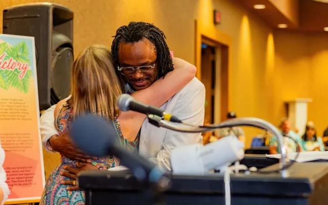 A man and woman hug warmly behind a podium in a conference room.