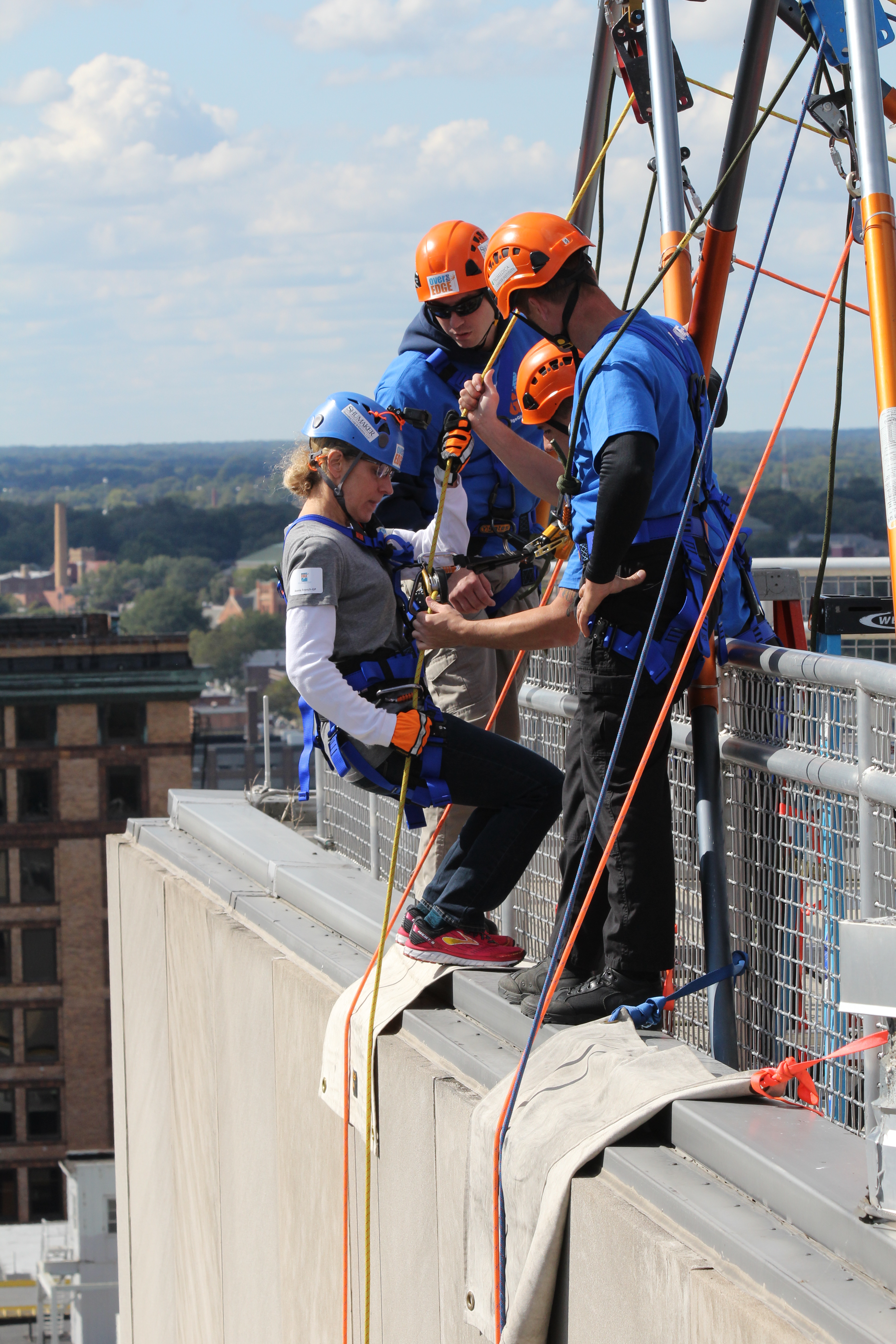 Three people in helmets and harnesses prepare to rappel down a building.