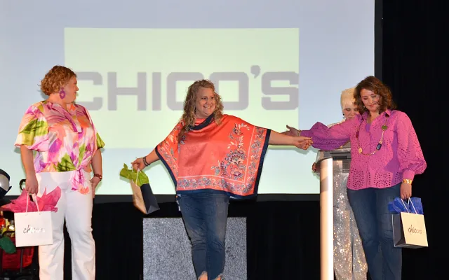 Three women on stage smiling, holding shopping bags.