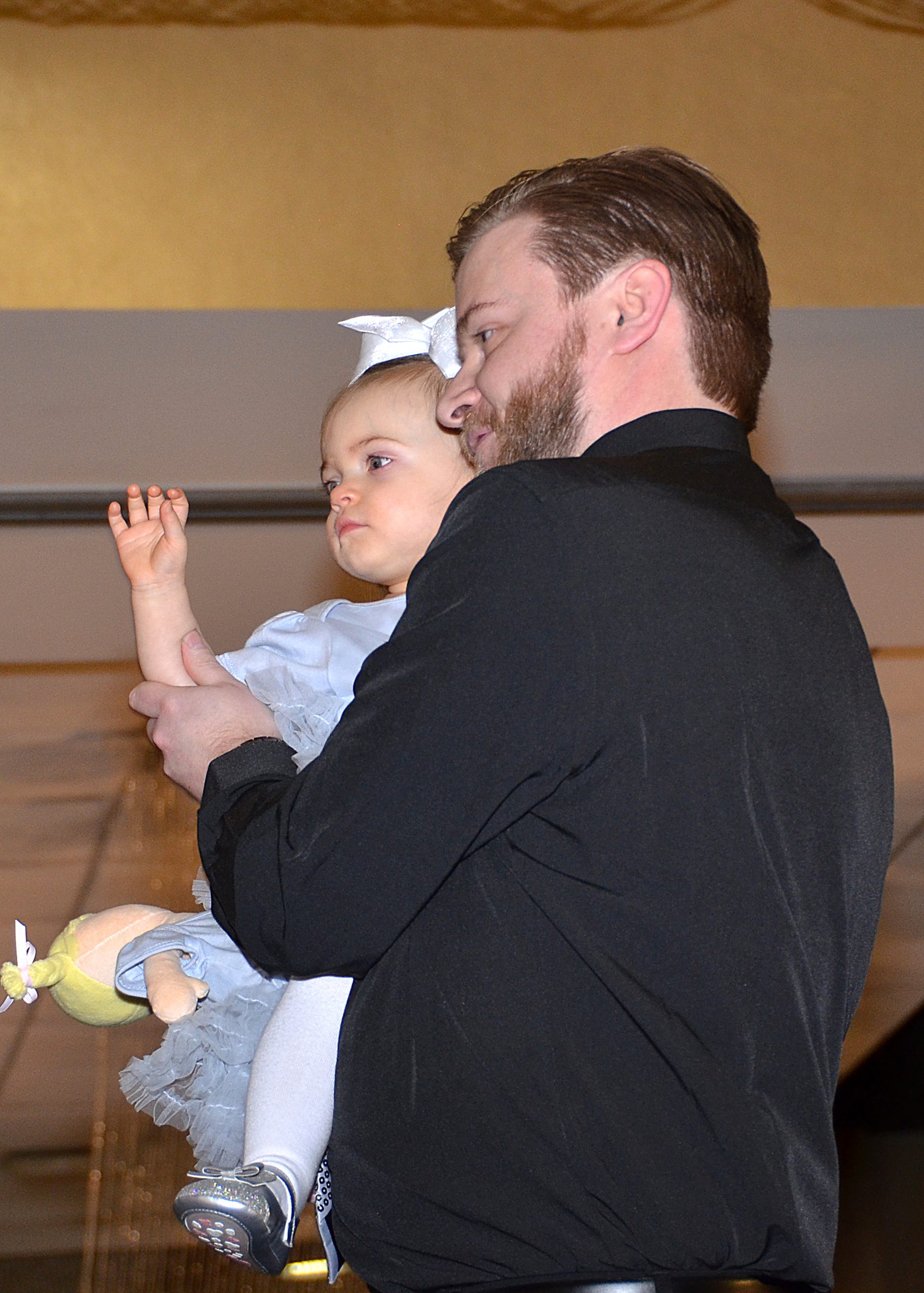 Man holding a baby with a bow, both gazing forward, indoors.