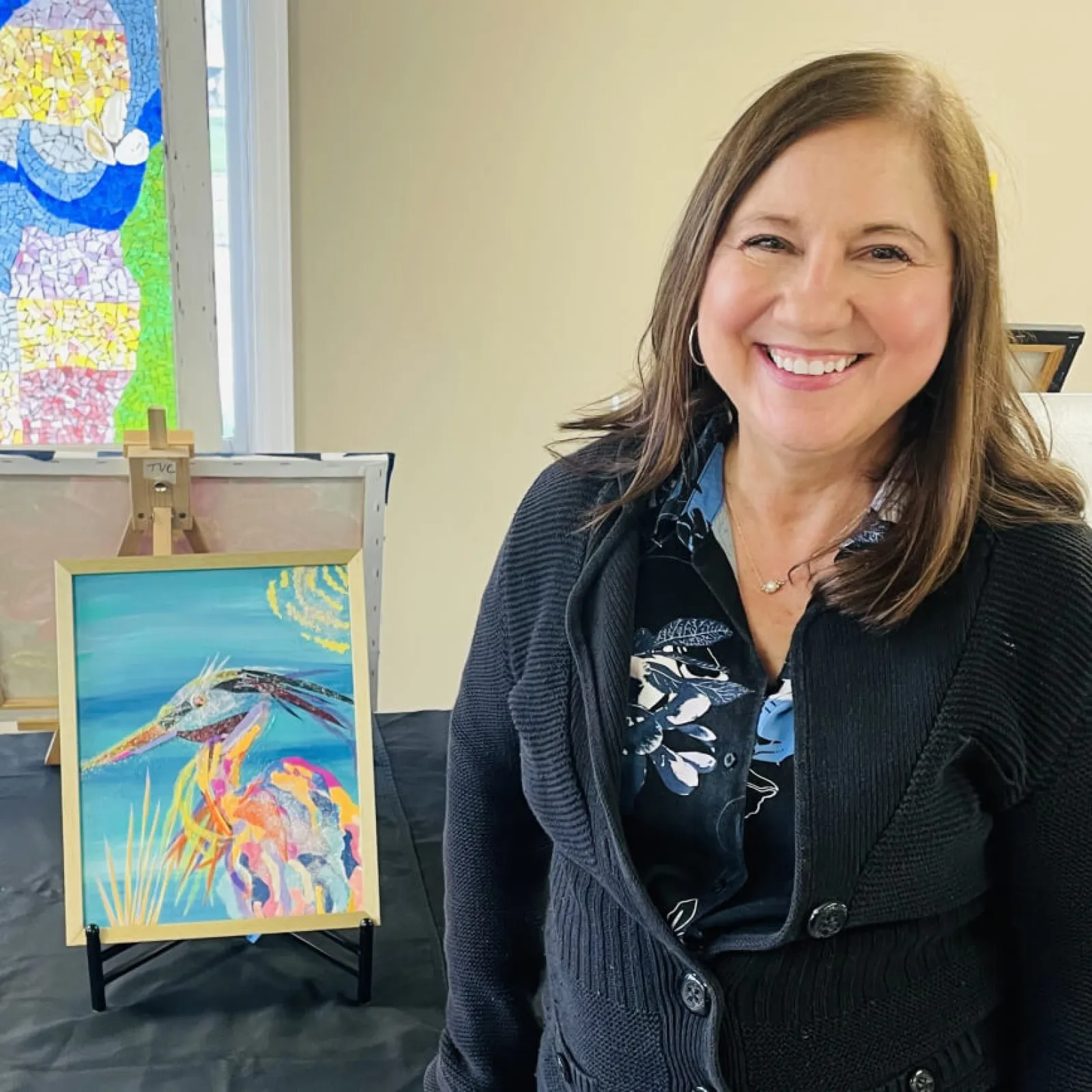 Smiling woman next to colorful painting on easel indoors.
