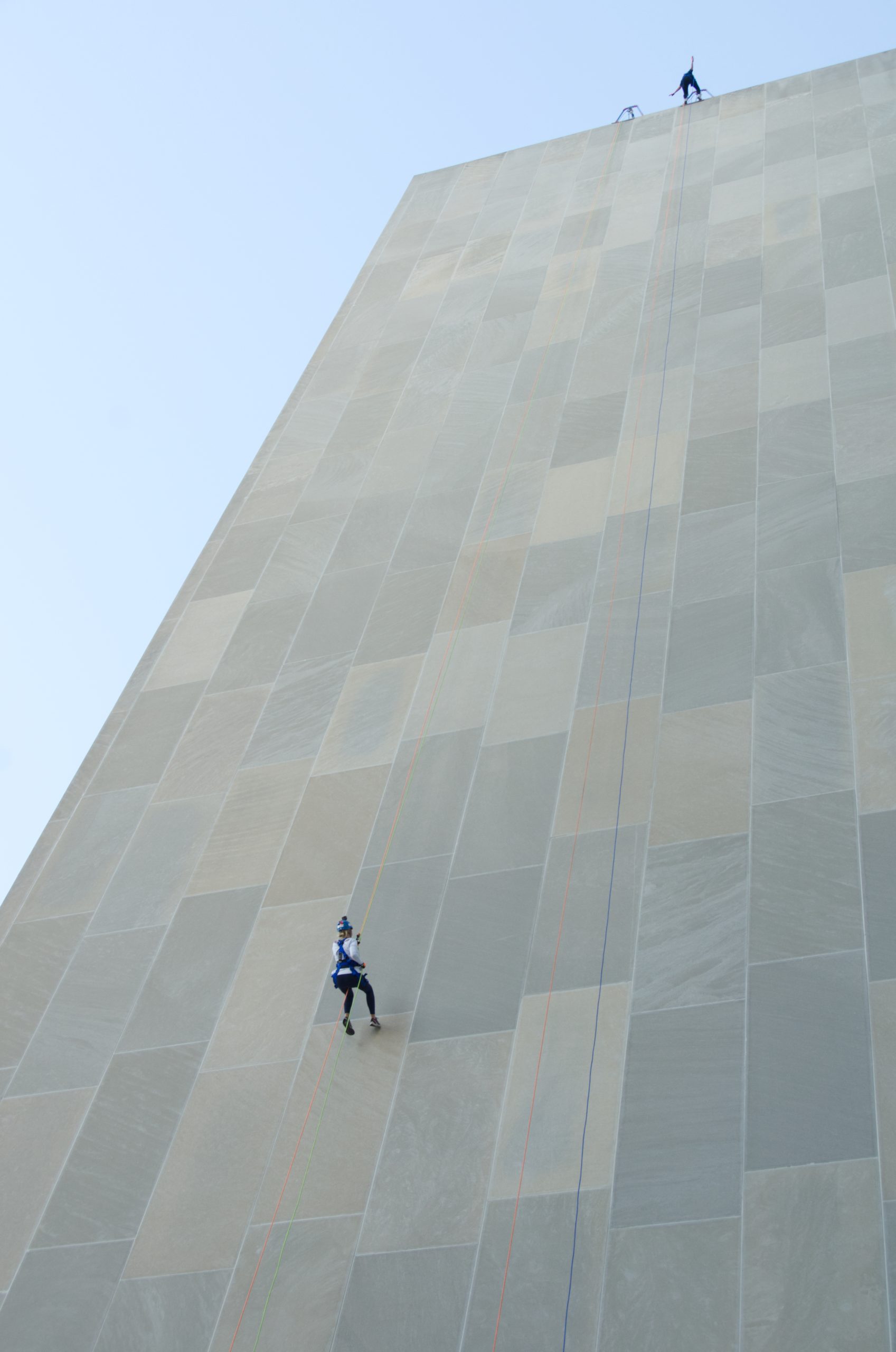 Two people rappelling down a tall, beige building wall.