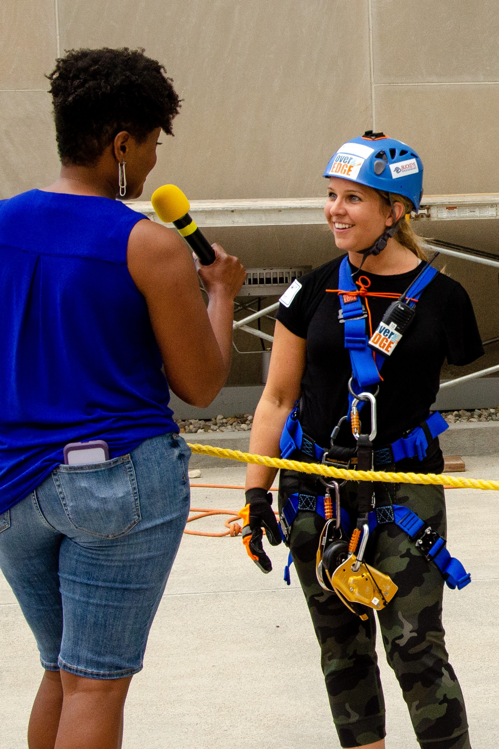 A woman in a blue helmet and harness being interviewed outdoors.