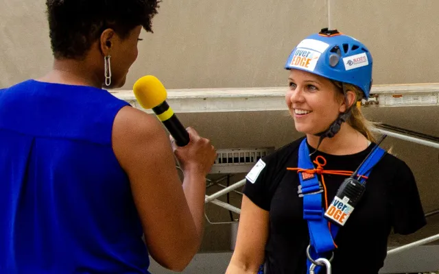 A woman in a blue helmet and harness being interviewed outdoors.