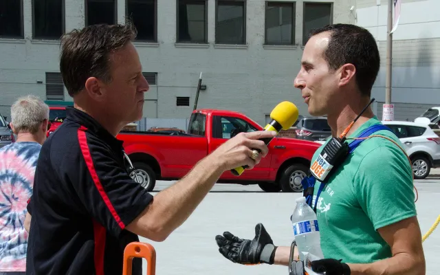 A man in a green shirt being interviewed by another holding a yellow microphone.