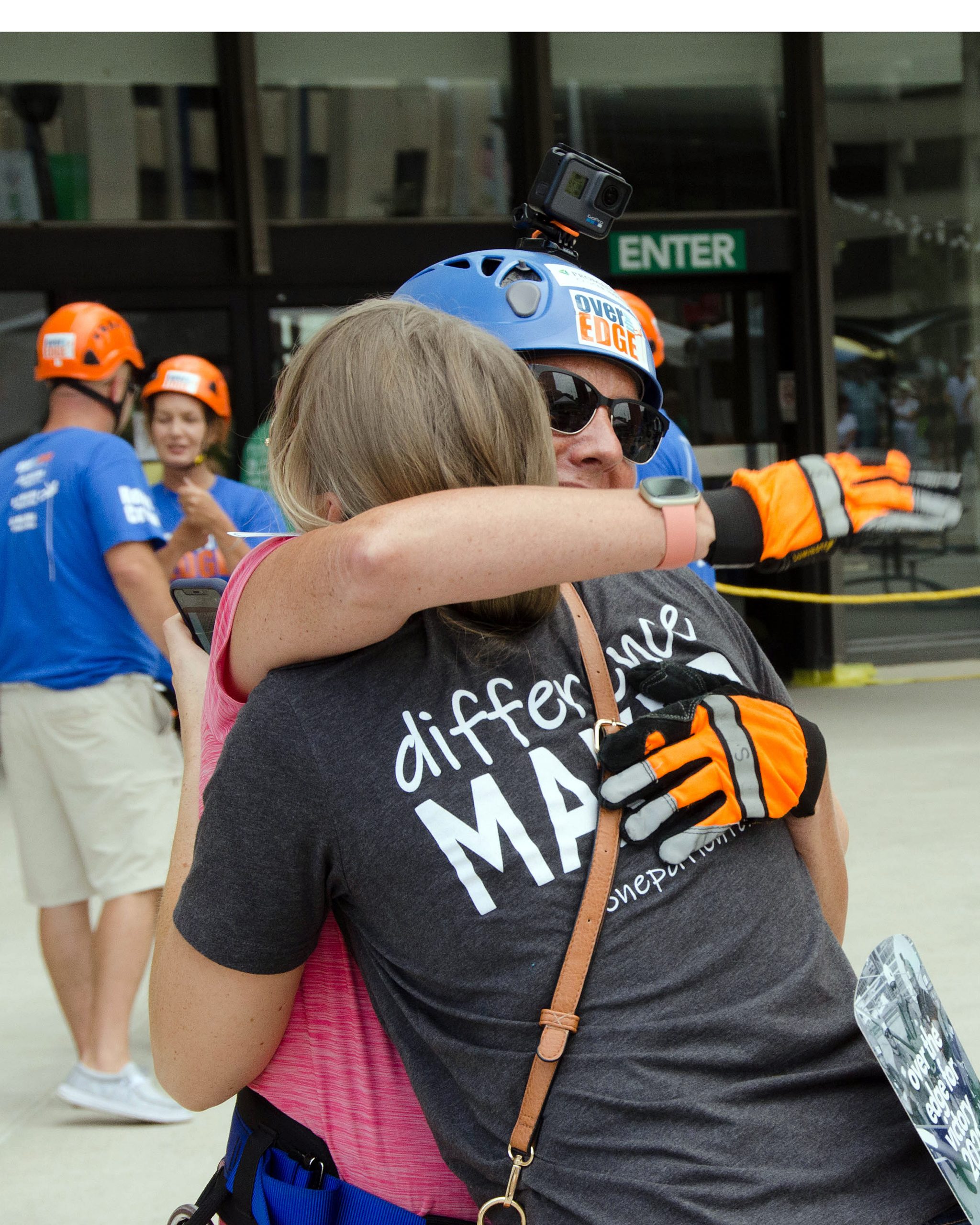 Two people wearing helmets hug outside, others with helmets in the background.