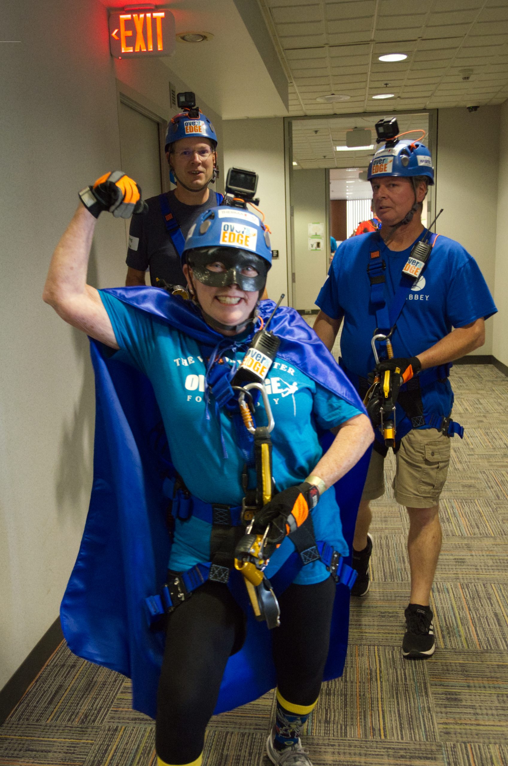 Three people in safety gear and blue attire in a hallway, one posing enthusiastically.