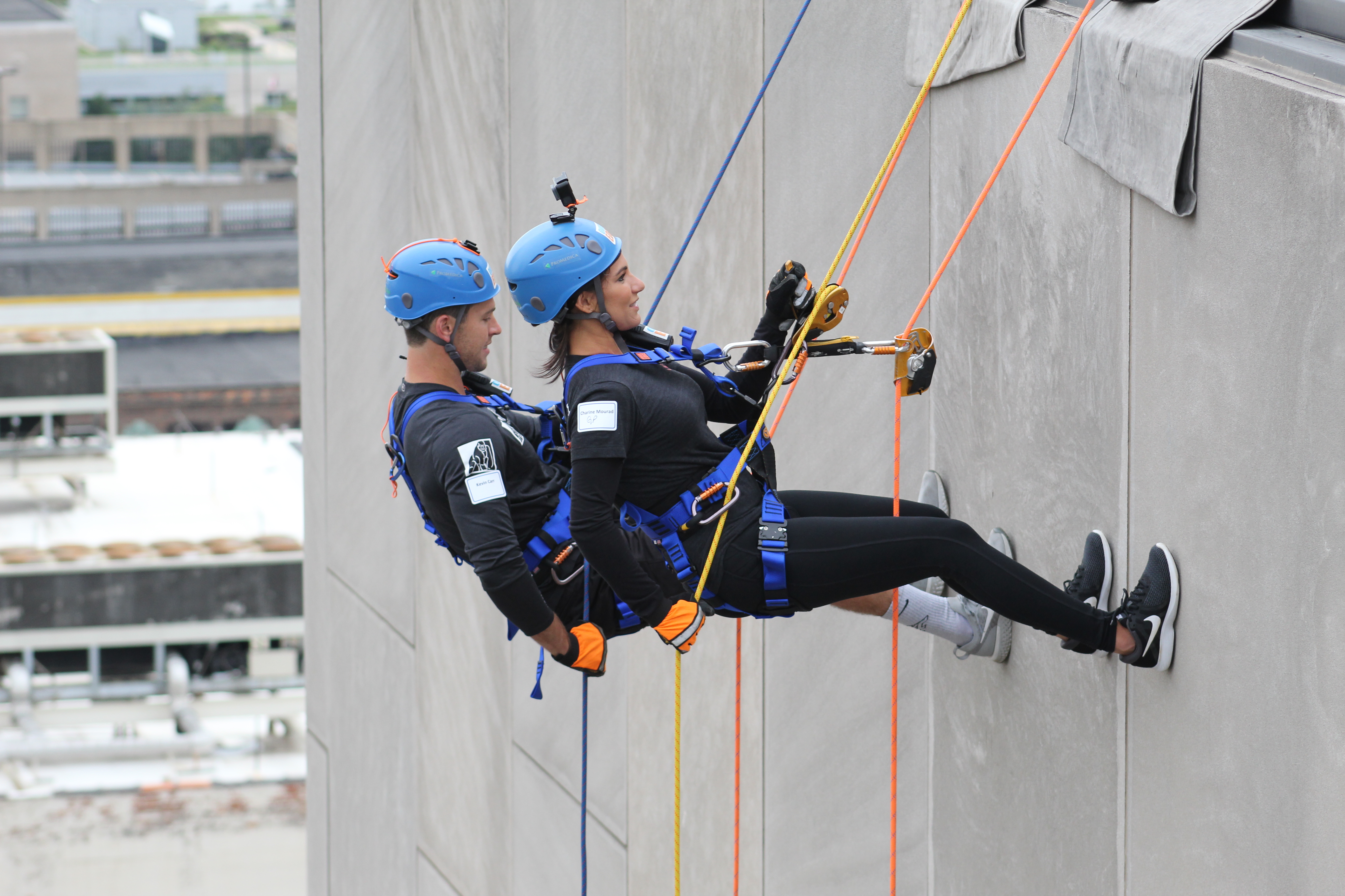 Two people in helmets and harnesses rappel down a building.