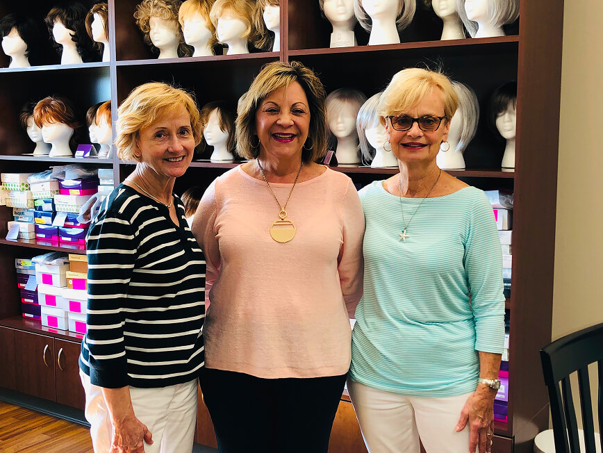 A group of women smiling with a collection of wigs behind them