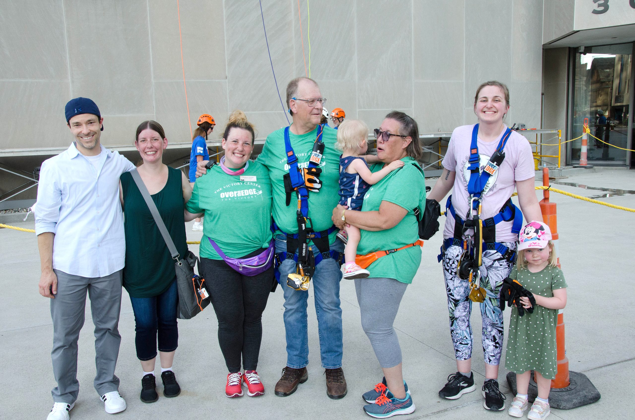 A group of smiling adults and children, some in climbing gear, standing together outdoors.