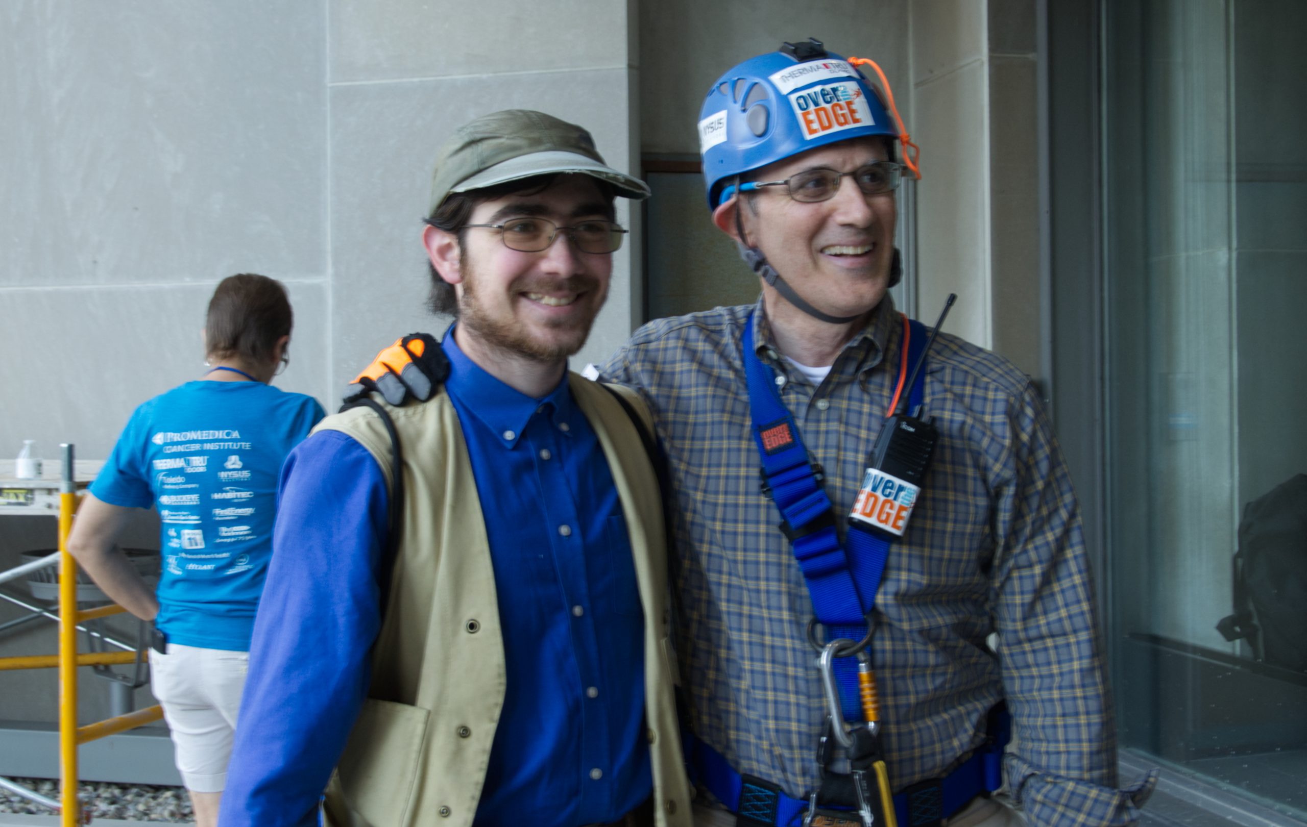 Two men smiling, one in climbing gear, the other in a hat, standing indoors.