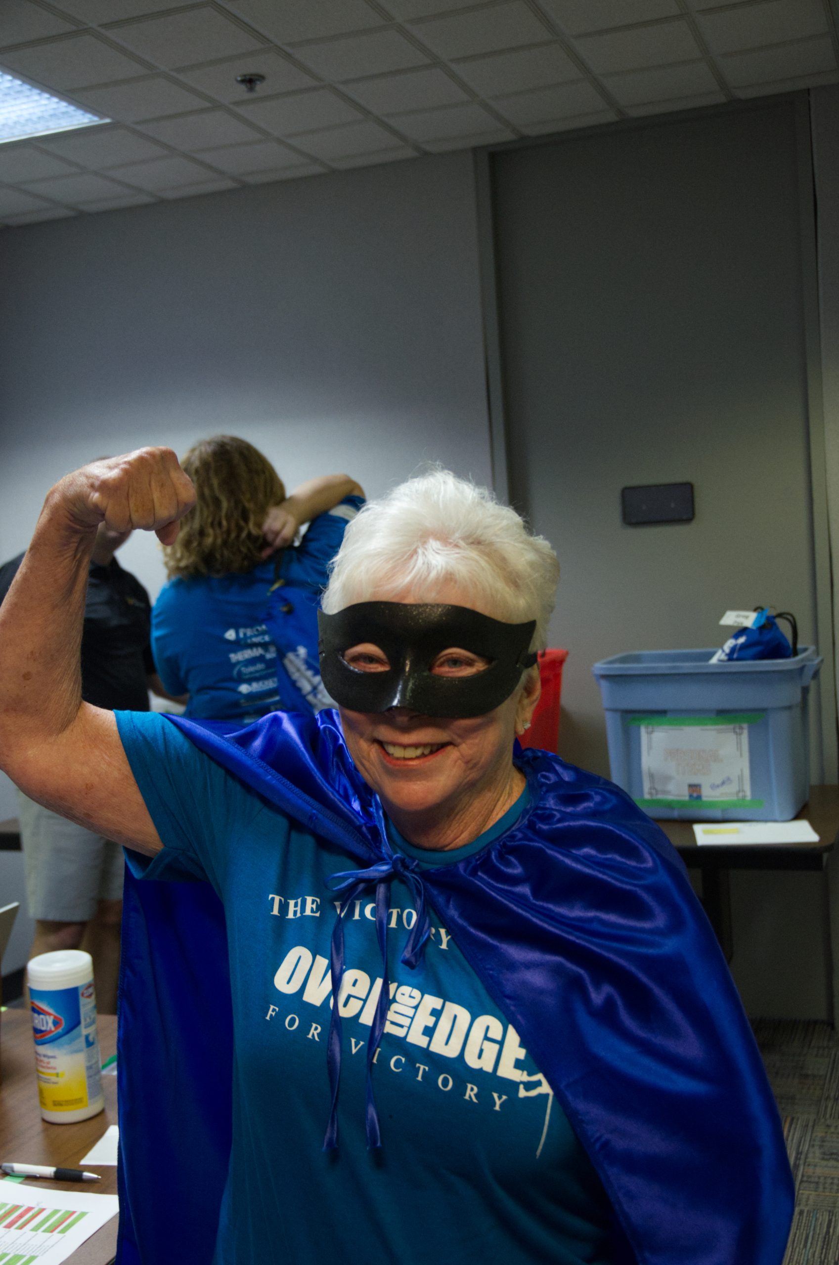 Person in blue cape and mask flexing arm confidently indoors.