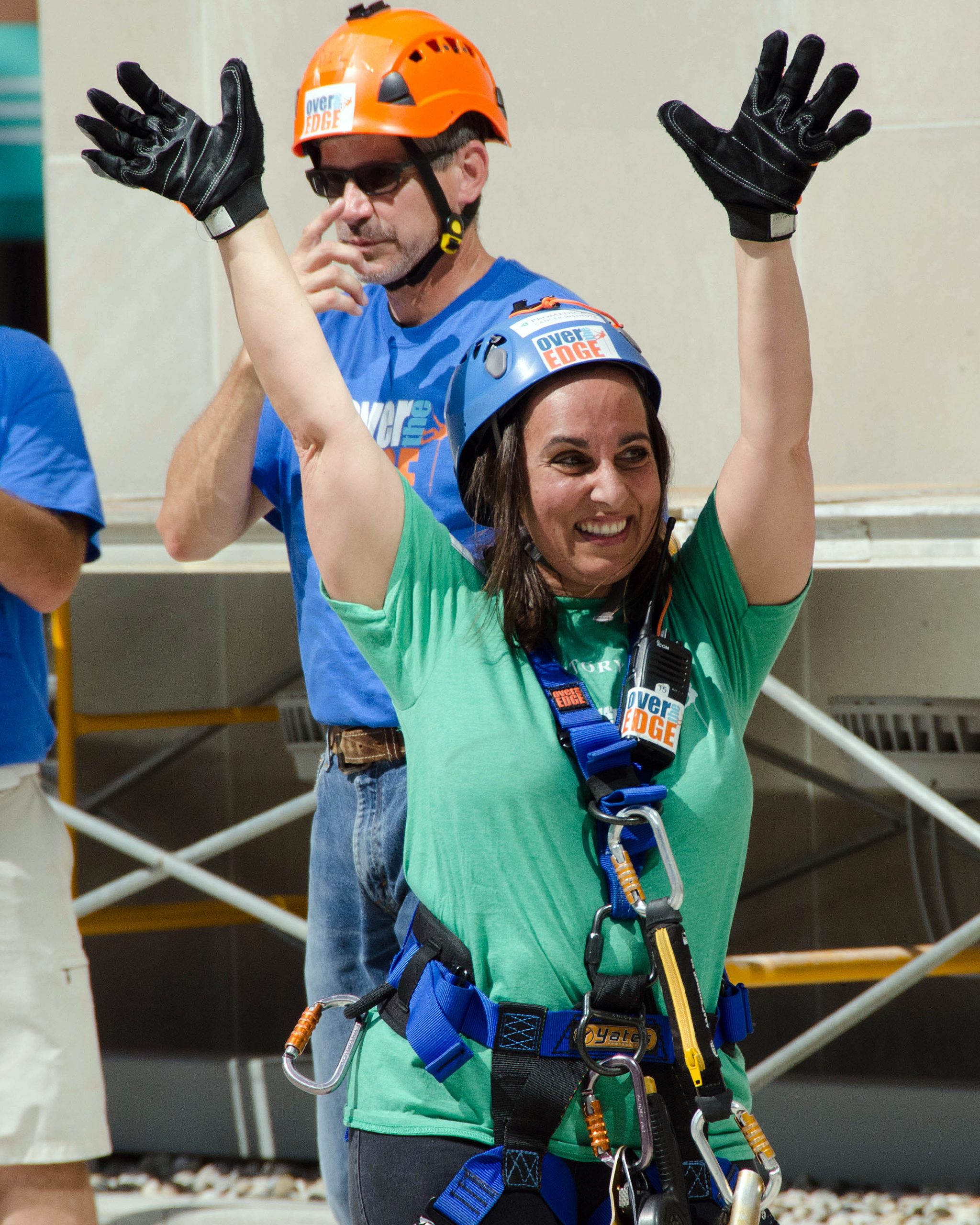 Woman in climbing gear excitedly raising arms, man in background wearing helmet.