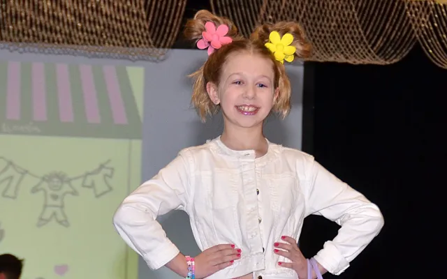 Young girl on runway, smiling, in colorful skirt and white top, with flowers in hair.