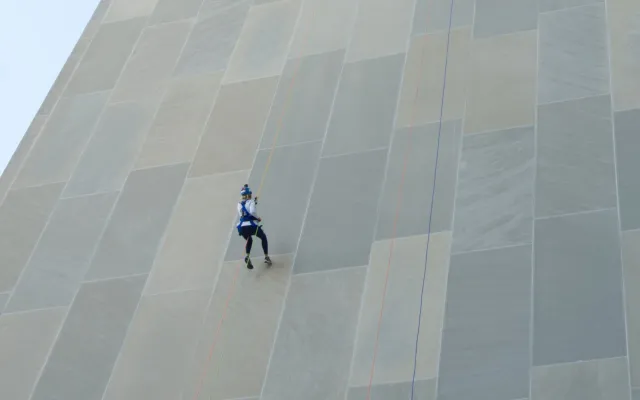 Two people rappelling down a tall, beige building wall.